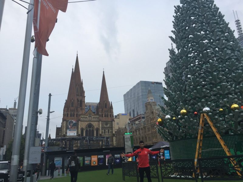 chico mirando a cámara y detrás los edificios de Melbourne y el árbol de Navidad con el cielo nublado